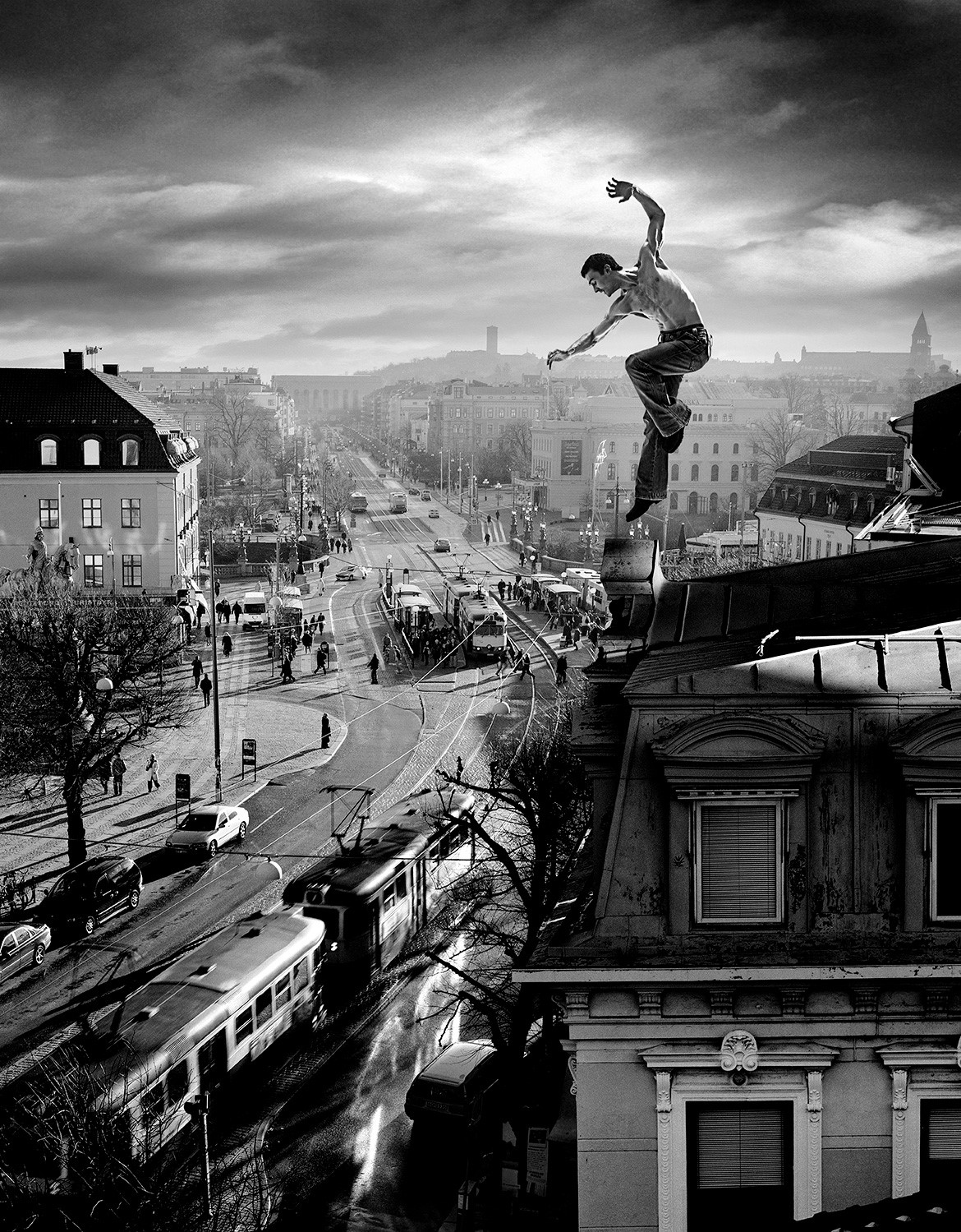 A person is jumping between two buildings against a backdrop of the Avenyn, Gothenburg. In black and white, the individual is captured in the air, just as they leap from one roof to another, creating a sense of motion and danger. The buildings have classical architecture with detailed patterns and structures. Below the buildings, a lively streetscape is visible with cars, trams, and pedestrians moving about. The city silhouette stretches beyond the buildings with various types of houses and trees visible; it appears to be a busy urban environment. The sky is filled with clouds that are backlit, producing an unearthly glow.