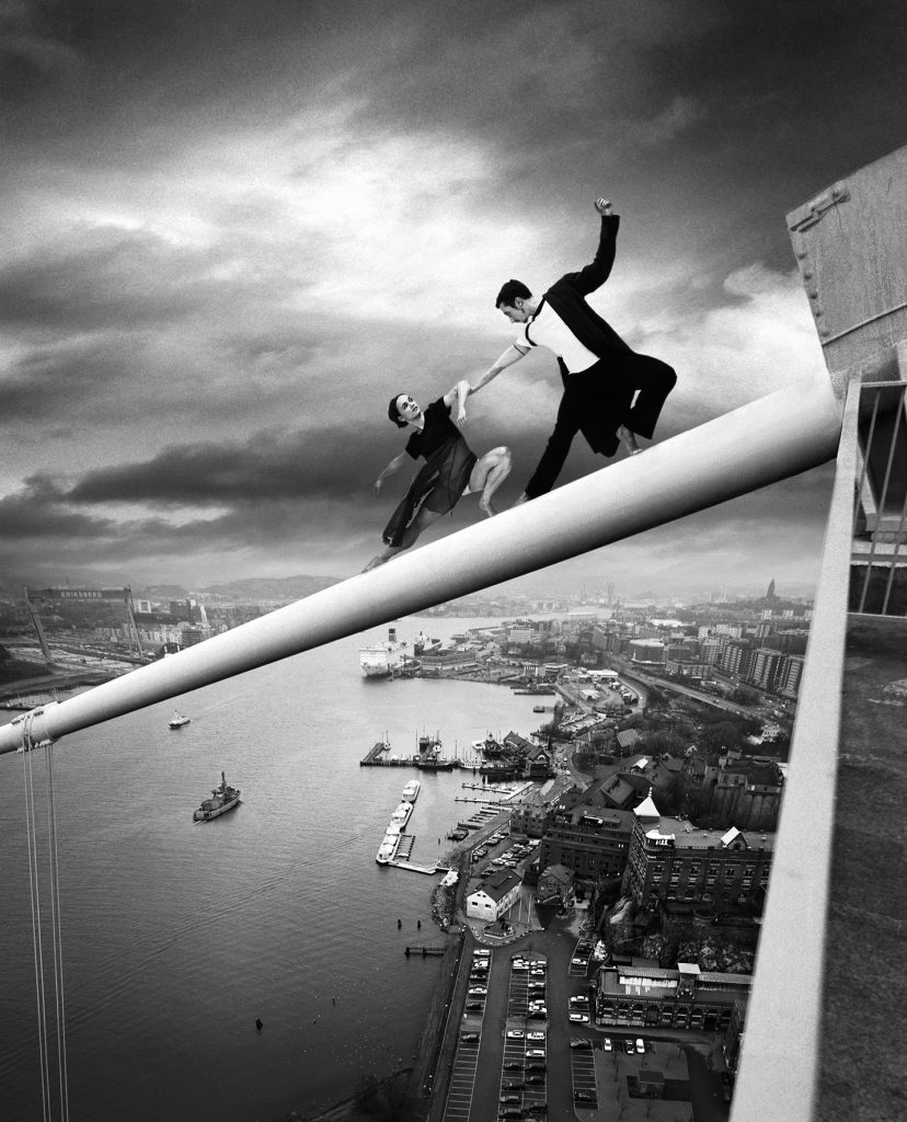 Two individuals are dancing on a beam that extends from the top of the Älvsborgsbron (Älvsborg Bridge). The person closest to the edge appears to be sliding or losing balance, while the other is reaching out to try to pull them back to safety. Below them is a sweeping view of Gothenburg with buildings, roads, and boats in the water. The sky is cloudy and threatening, adding to the intense mood of the moment.